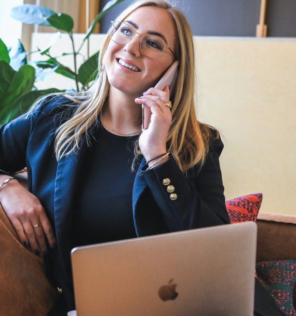Junge Frau im Kundengespräch am Telefon mit Laptop auf dem Schoß im modernen Büro
